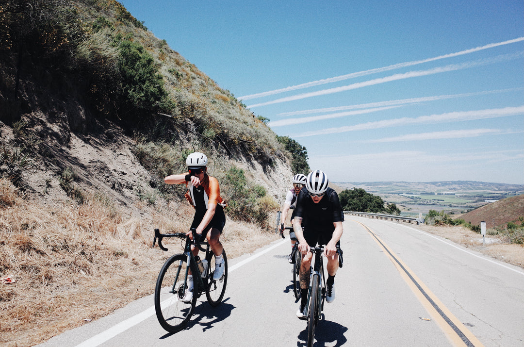 three cyclists riding uphill towards the viewer on a street, light blue sky in the background